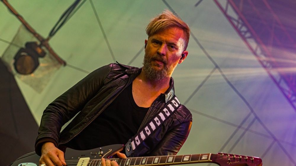 Organek na scenie, gra na gitarze, patrzy prosto w obiektyw. Ma na sobie skórzaną czarną kurtkę.