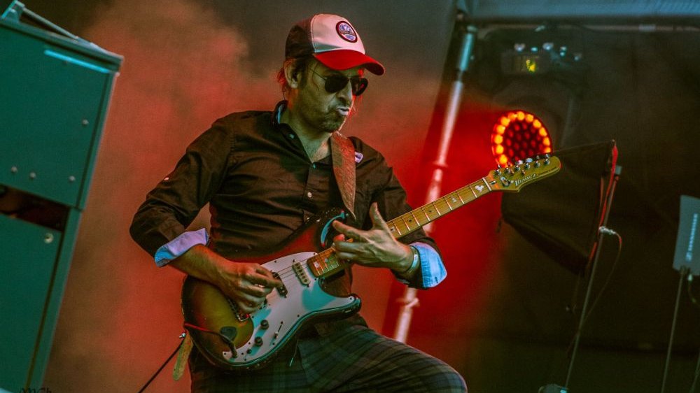 Na scenie gitarzysta, grający na gitarze elektrycznej, ma na sobie czarne okulary i wzorzystą czapkę z daszkiem.