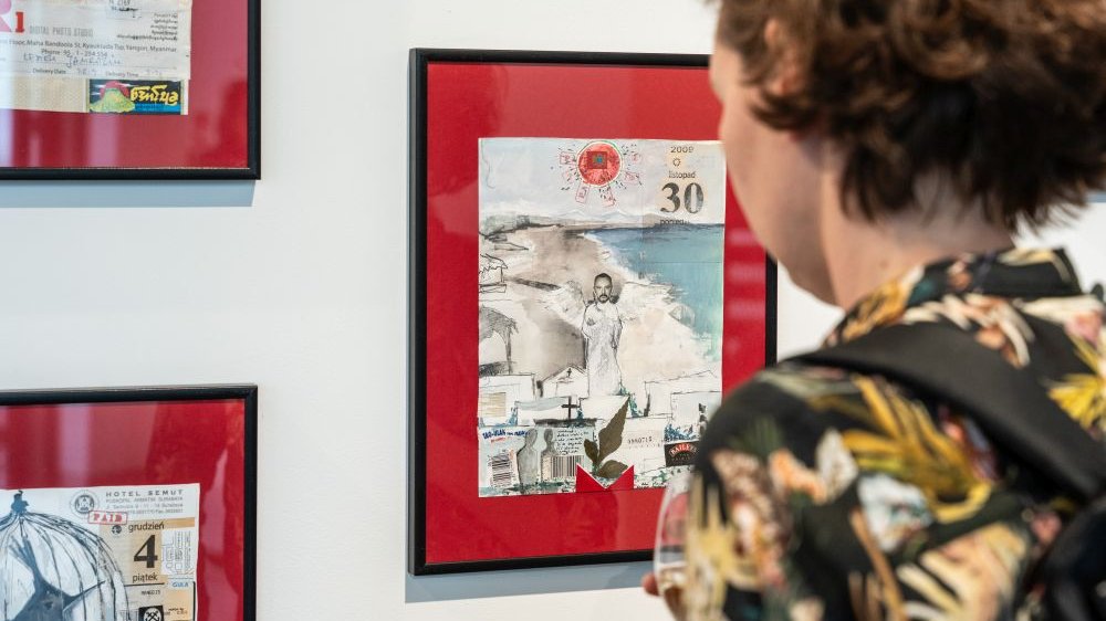 Stojąca tyłem do aparatu dziewczyna ogląda grafiki na ścianie galerii. To małe kolaże umieszczone w ramkach na czerwonym tle.