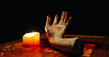 Zakrwawiona i wytatuowana zabalsamowana ludzka ręka leży na stole w nienaturalnej pozie. Obok niej płonąca świeca i zużyte zapałki.