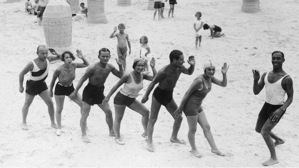 Czarno-białe zdjęcie kilku osób w letnich strojach w tej samej postawie - z podniesioną ręką i wystawioną do przodu nogą.