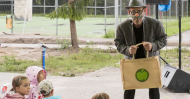 Zabawny wąsaty pan w szarym meloniku i okrągłych okularach uśmiecha się do dzieci na widowni, niosąc w obu rękach ekologicznę torbę z narysowanym zielonym jabłkiem.