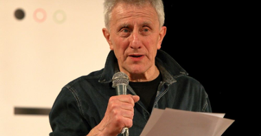 Poeta jest ubrany w ciemną, dżinsową kurtkę, w ręku trzyma mikrofon oraz plik kartek. Opowiada coś prawdopodobnie na scenie.