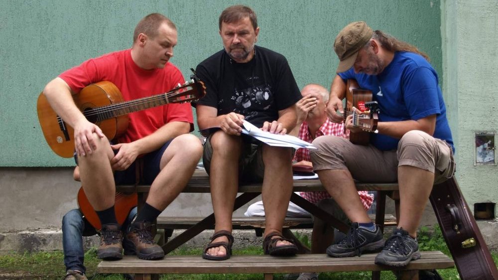 Trzech mężczyzn siedzi obok siebie na drewnianym stole, są w trakcie tworzenia muzyki, dwóch trzyma gitarę, jeden, w środku, zbiór kartek, prawdopodobnie nut.