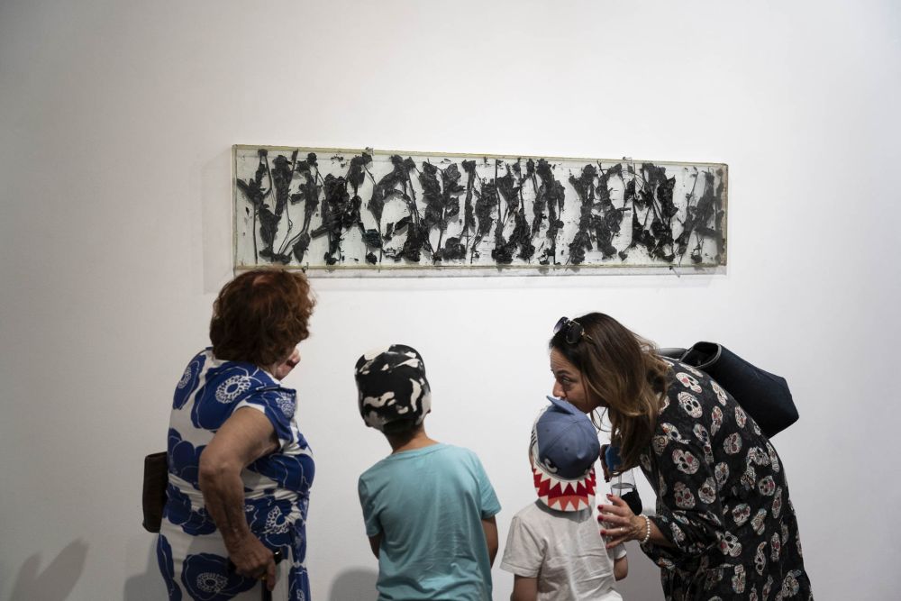 Dwie obserwatorki wraz z dziećmi przyglądają sie wywieszonemu na białej ścianie dziełu sztuki autorstwa artysty. - grafika artykułu
