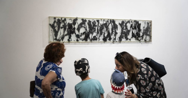 Dwie obserwatorki wraz z dziećmi przyglądają sie wywieszonemu na białej ścianie dziełu sztuki autorstwa artysty.