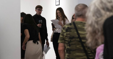 Ludzie w różnym wieku zwiedzają galerię podczas wernisażu, niektórzy trzymają w ręku kieliszek wina, inni czytają opis kuratorski wystawy. Na ścianie za uczestnikami wisi oprawione w ramkę hasło "Learn".