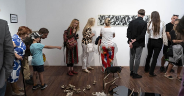 Grupa ludzi zwiedza wystawę podczas wernisażu, rozglądając się po galerii. Na podłodze leży kompozycja z kości i czaszki z porożem, na ścianie wisi czarno-białe dzieło sztuki.