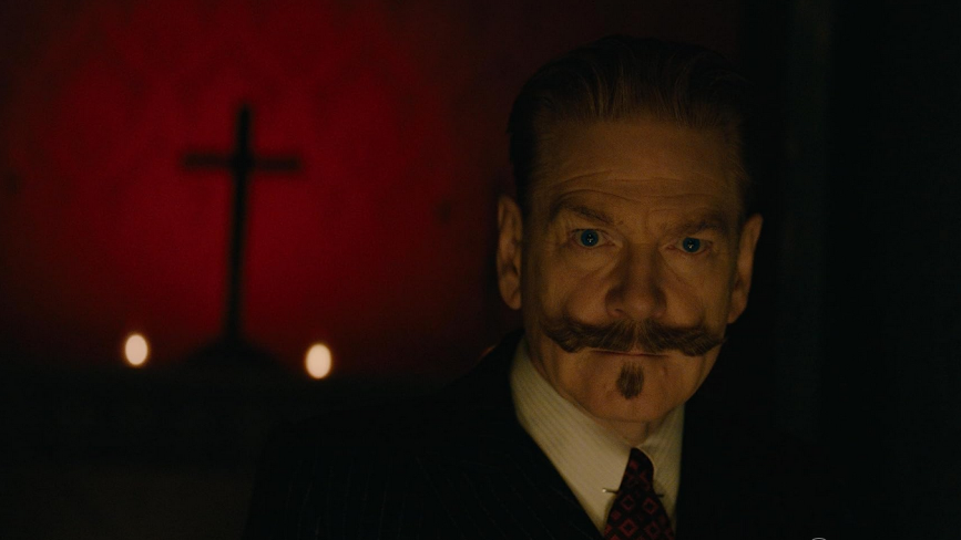 Hercules Poirot patrzy uważnie w kierunku widza. Ma bardzo wyrazisty, podkręcony wąs i elegancki garnitur. Za nim widać oświetloną na czerwono ścianę, na której wisi krzyż. - grafika artykułu