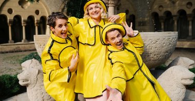 Trójka aktorów w żółtych pelerynach, kapeluszach i kaloszach przeciwdeszczowych uśmiacha się szeroko do obiektywu.