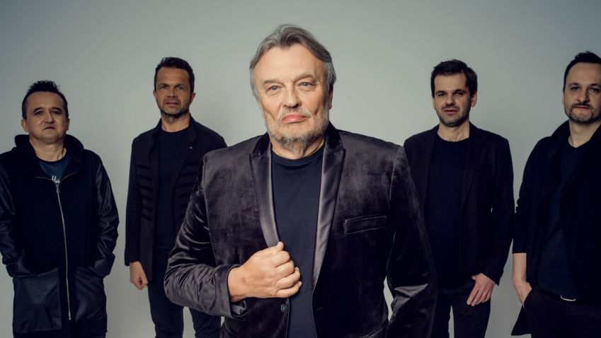 Krzysztof Cugowski jest ubrany w czarny garnitur z aksamitu, czarną koszulkę oraz spodnie. Za nim stoi czterech członków zespołu - wszyscy w czerni. - grafika artykułu