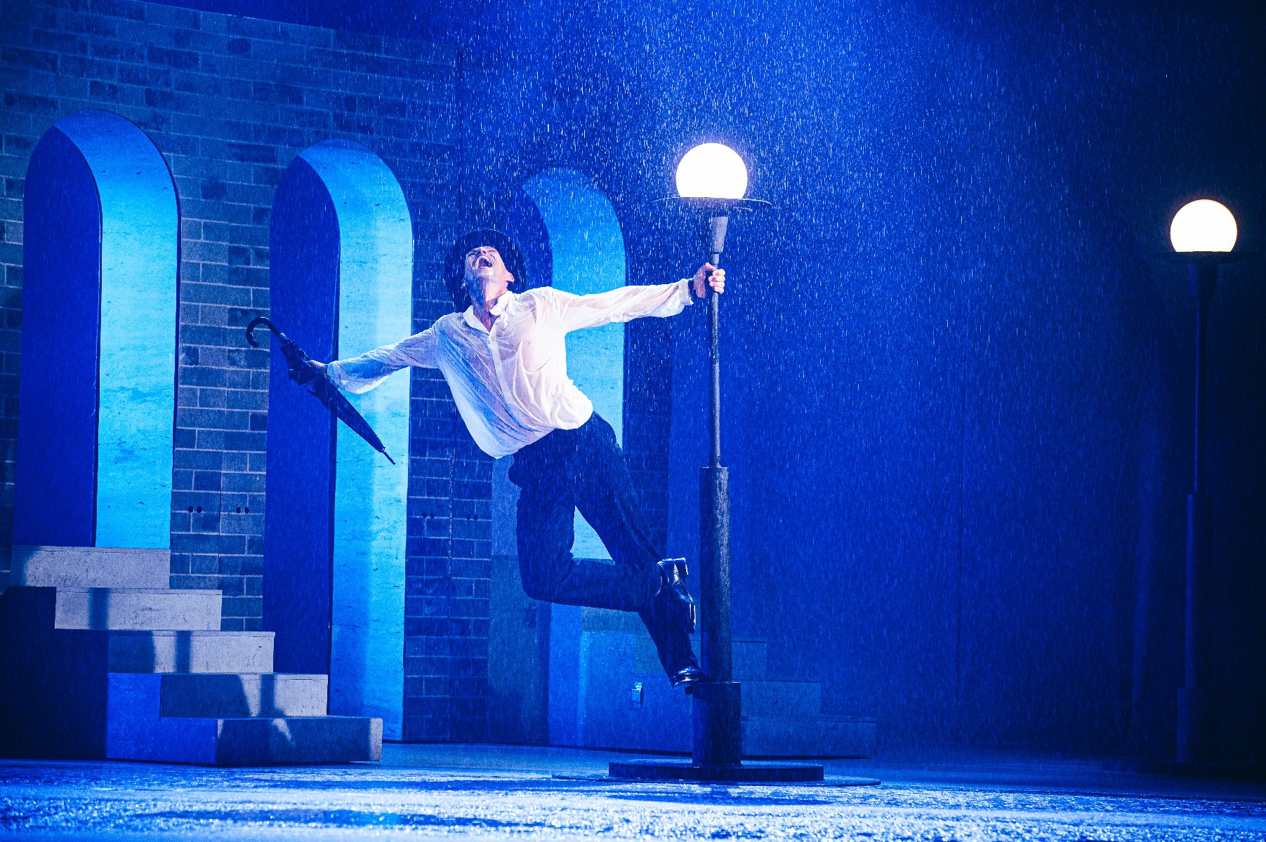Aktor odtwarza na scenie ikoniczną scenę z Deszczowej Piosenki, tańczy stojąć na postumencie latarni ulicznej - grafika artykułu