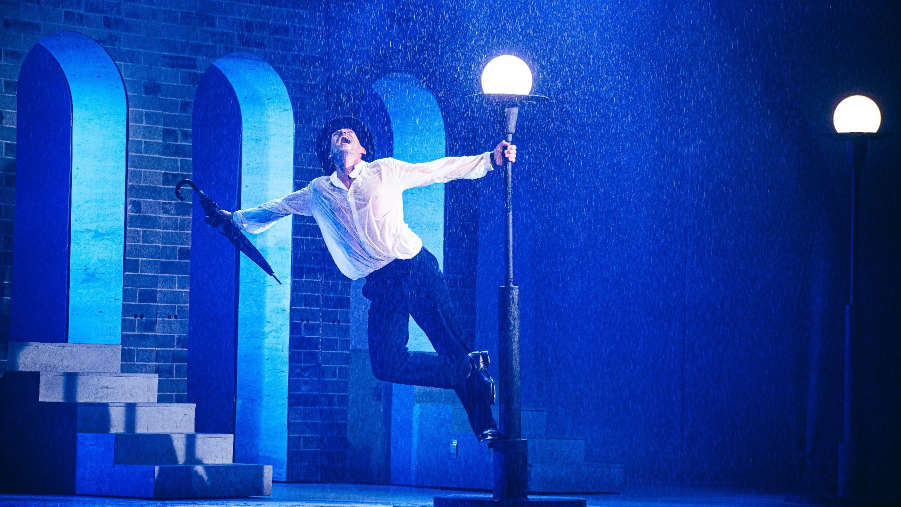 Aktor odtwarza na scenie ikoniczną scenę z Deszczowej Piosenki, tańczy stojąć na postumencie latarni ulicznej