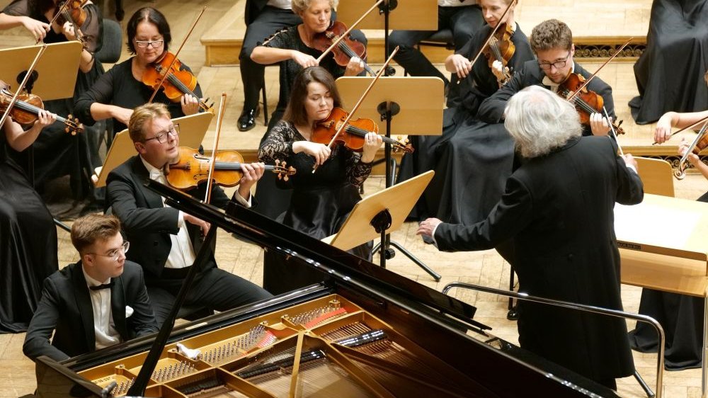 Pianista spogląda na orkiestrę dyrygowaną przez siwego mężczyznę.