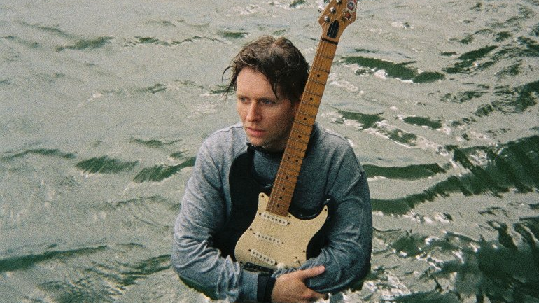 Mężczyzna trzyma gitarę i stoi do pasa w wodzie.