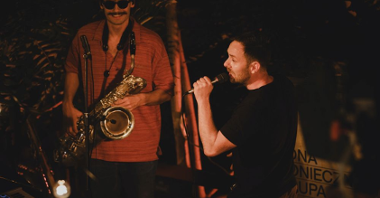 Łona w czarnej koszulce stoi na scenie i rapuje z mikrofonem w dłoni, obok niego stoi uśmiechnięty saksofonista w pomarańczowej koszulce.