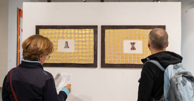 Dwoje zwiedzających ogląda oprawione w "wyszyte" ramy tkaniny, na których ktoś naszył złote kółka, a na środku widnieje naszyty obrazek.