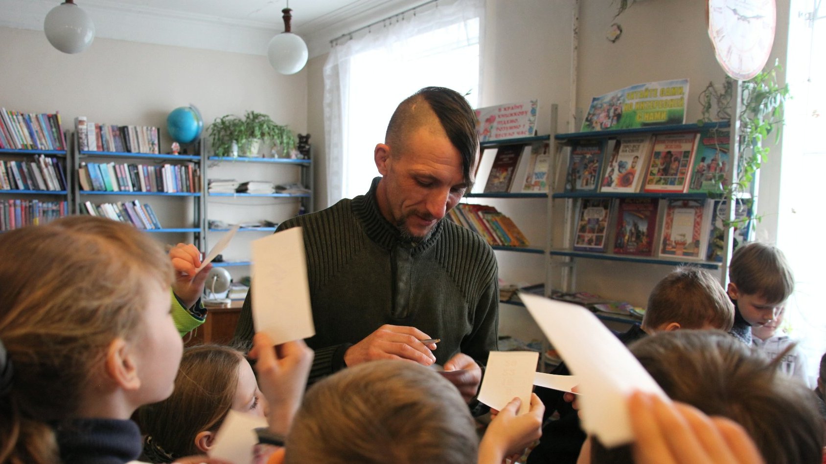 W centrum mężczyzna z długopisem w dłoni. Wokół dzieci z kartkami papieru.