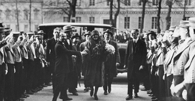 Kobieta w futrze wychodzi z samochodu w towarzystwie kilku mężczyzna. Po prawej i lewej stronie rząd osób z podniesionymi rękami.