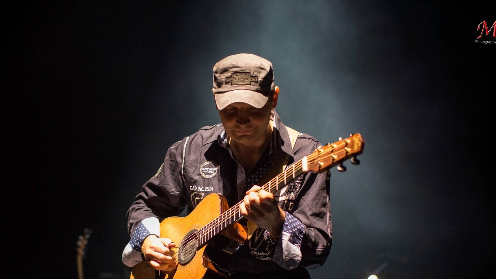 Mężczyzna w czapce gra na scenie, trzyma w rękach gitarę, ma spuszczony wzrok.