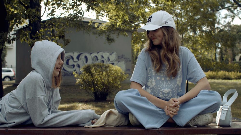 Lato, dwie młode, nastoletnie dziewczynki siedza na murku. Obie są ubrane sportowo, mają na sobie koszulki, bluzy, czapki z daszkiem, obie się uśmiechają. - grafika artykułu