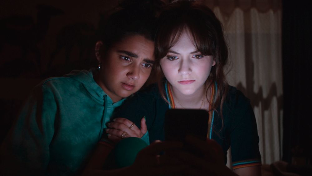 Dwie młode dziewczyny, biała i ciemnoskóra, oglądają coś na ekranie komórki, siedząc w ciemności. - grafika artykułu