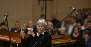 Agnieszka Duczmal dyryguje orkiestrą, ma zamknięte oczy. Za nią zasłuchana publiczność.