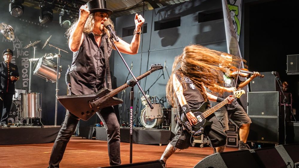 Koncert metalowego zespołu. Wokalista w cylindrze na głowie i basista z rozwianymi włosami wykonują energiczne ruchy.