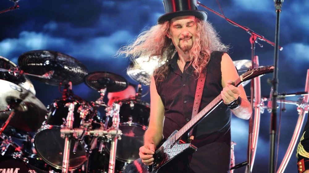 Długowłosy gitarzysta gra na gitarze elektrycznej, na głowie ma czarny cylinder.