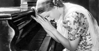 Czarno-biały kadr filmowy. Kobieta opiera się o wieko fortepianu.