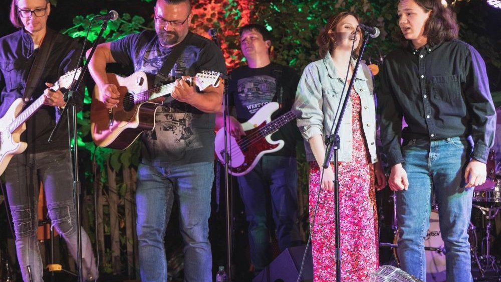 Muzycy występują na scenie, trzech mężczyzn gra na gitarach, młodzież śpiewa.