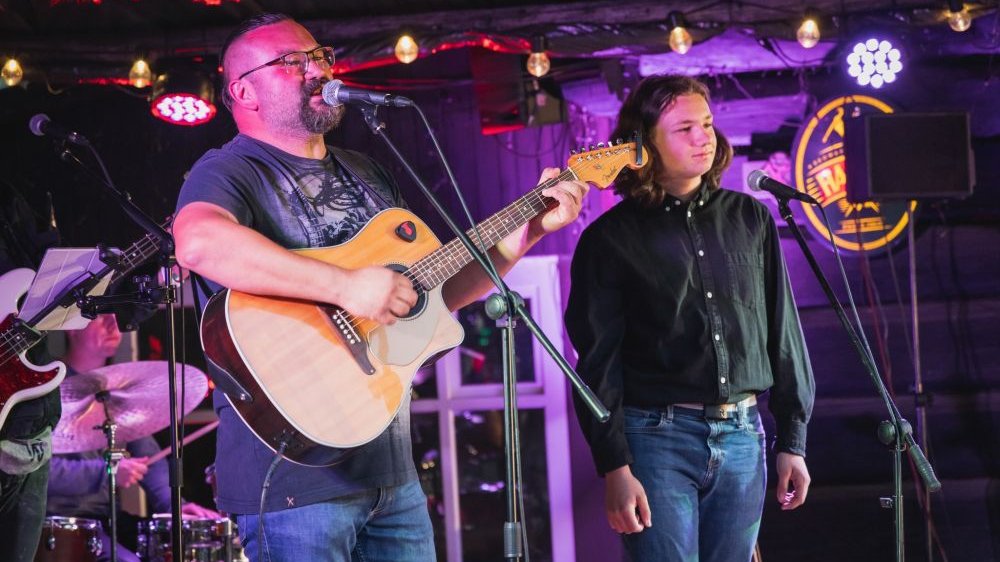 Koncert zespołu, lider z gitarą akustyczną przy mikrofonie, obok niego młody chłopak.