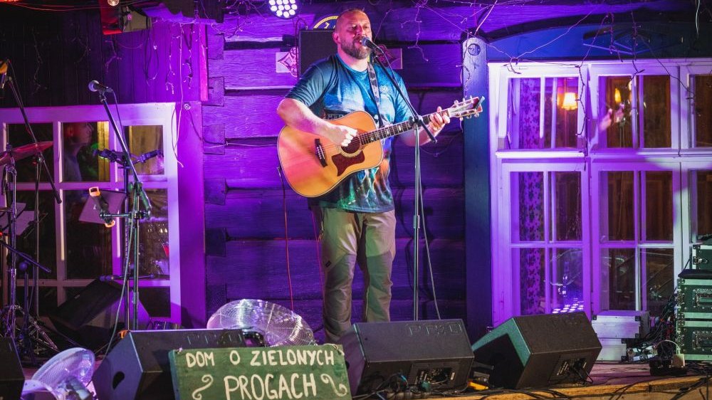 Meżczyzna z gitarą śpiewa na scenie. Za nim dekoracje przypominające stary dom.