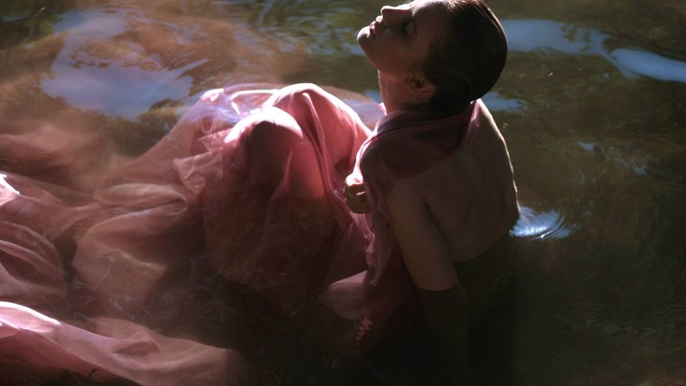Kobieta w długiej sukni z odsłoniętymi plecami siedzi w wodzie, twarz unosi ku niebu. - grafika artykułu