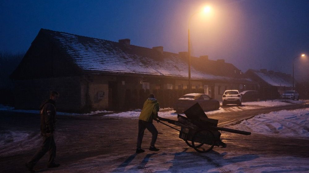 Dwóch mężczyzn idzie zaśnieżoną drogą po zmroku, w świetle latarni. Jeden z nich pcha taczkę z opałem.