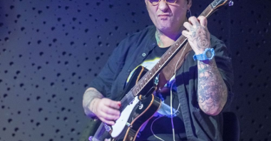 Mężczyzna w okularach i kapeluszu gra na gitarze elektrycznej.