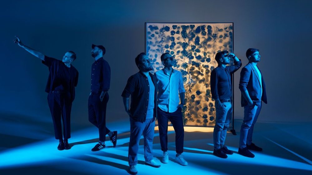 Sześciu mężczyzn stoi w niebiesko oświetlonej sali ze złotą ścianą z malunkami w kształcie liści. - grafika artykułu