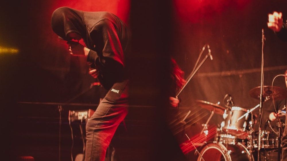 Raper w czarnej bluzie z kapturem występuje na scenie zgięty w pół. Za jego plecami perkusja, dookoła czerwone światło.