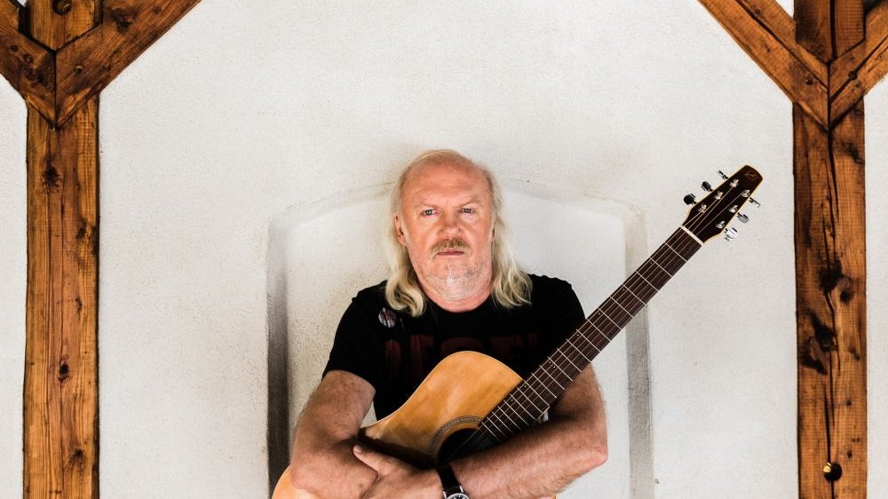 Muzyk z gitarą na tle białej ściany. Po prawej i lewej stronie drewniane filary.