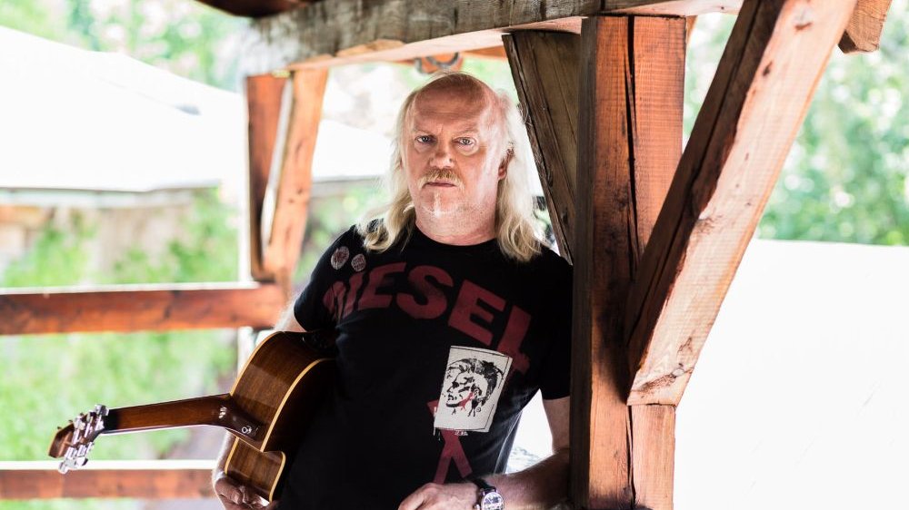 Artysta stoi na drewnianej werandzie z gitarą pod pachą. Za nim rozmyta zieleń drzew.