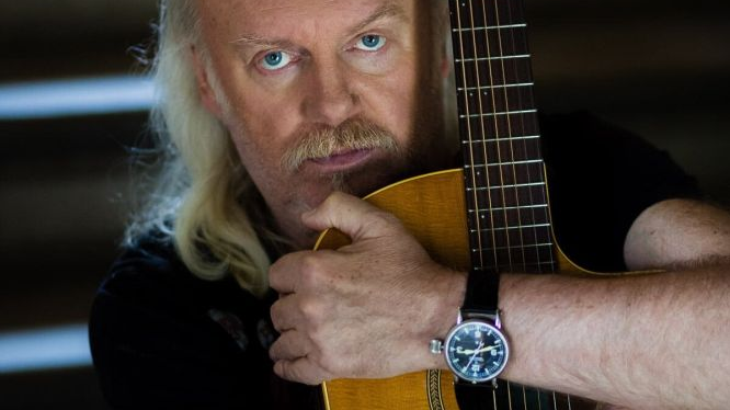 Mężczyzna w długich blond włosach i z wąsem obejmuje gitarę, patrzy prosto w obiektyw.