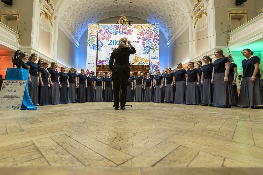 Występ chóru żeńskiego ustawionego w półokręgu na scenie Auli UAM pod przewodnictwem dyrygentki. - grafika artykułu