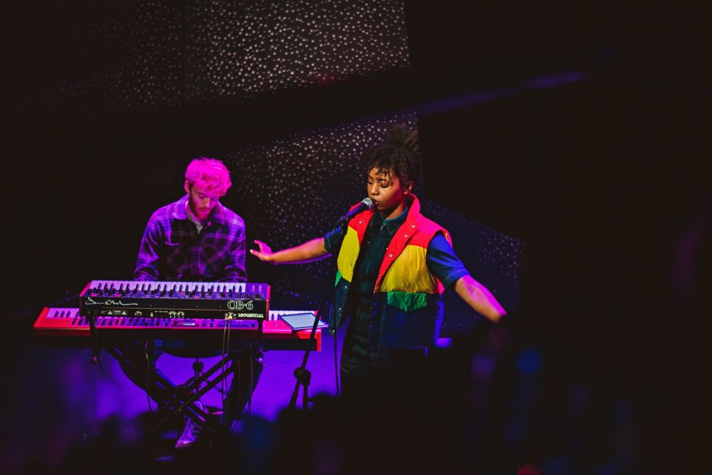 Wokalistka w kolorowej kamizelce śpiewa do mikrofonu, za nią meżczyzna za elektronicznym pianinem. - grafika artykułu
