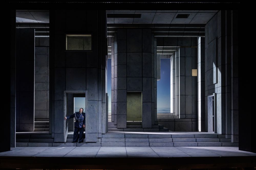 Surowa scenografia imitująca monumentalny budynek. W drzwiach stoi jeden aktor w wojskowym mundurze. - grafika artykułu
