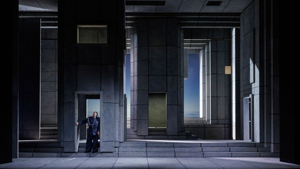 Surowa scenografia imitująca monumentalny budynek. W drzwiach stoi jeden aktor w wojskowym mundurze.