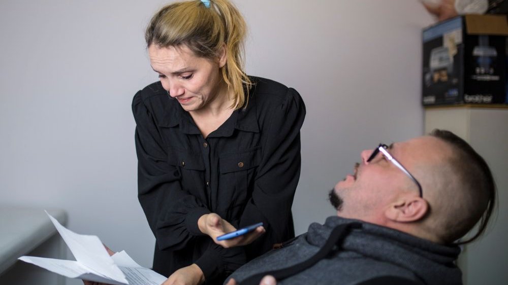 Kobieta w kucyku na głowie stoi, ma w ręce telefon komórkowy. Mężczyzna siedzi, przechyla się na krześle w tył. Oboje trzymają plik kartek.