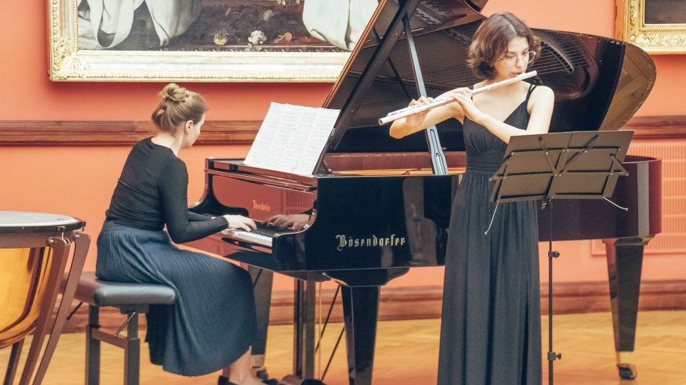Dwie kobiety grają na instrumentach, fortepianie i flecie. Za nimi pomarańczowe ściany muzeum, na nich wiszące obrazy.