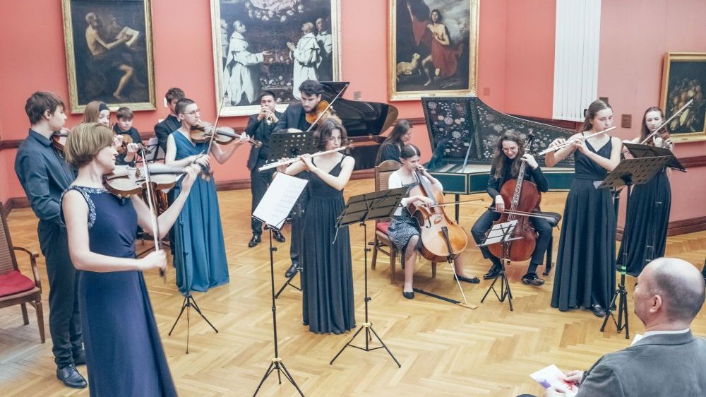 Młodzi muzycy grają na instrumentach, stoją lub siedzą blisko siebie.
