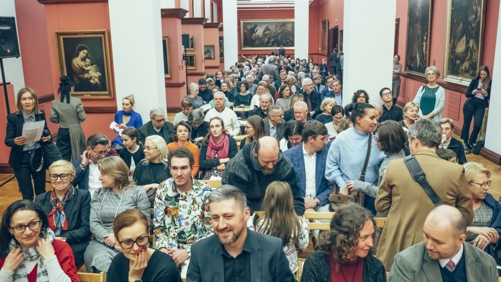 Liczna publiczność słucha koncertu siedząc. Dookoła nich obrazy na pomarańczowych ścianach.