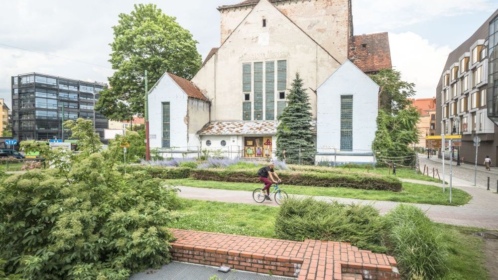 Mężczyzna jadący na rowerze po ścieżce. Za nim szara bryła synagogi.